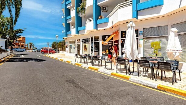 A street view with a row of outdoor seating, a building with blue and white exterior, and a clear sky. - Home Holidays 204 (CN)