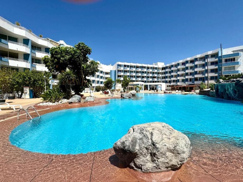 A hotel with a large swimming pool, multiple balconies, and a tropical landscape.