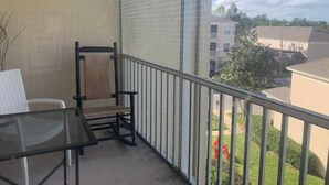 A balcony with a glass table and chairs, a metal railing, and a view of buildings and greenery. - Beautiful 2-bedroom condo in a serene gated community with refreshing AC (Kissimmee)