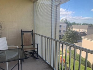 A balcony with a glass table and chairs, a metal railing, and a view of buildings and greenery. - Beautiful 2-bedroom condo in a serene gated community with refreshing AC (Kissimmee)