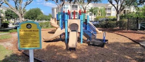 Un parque infantil con un tobogán y estructuras para escalar, rodeado de árboles y un edificio al fondo.