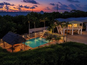 Ein Poolbereich mit einer strohgedeckten Struktur, einer überdachten Sitzgelegenheit und Blick auf den Sonnenuntergang über dem Meer.