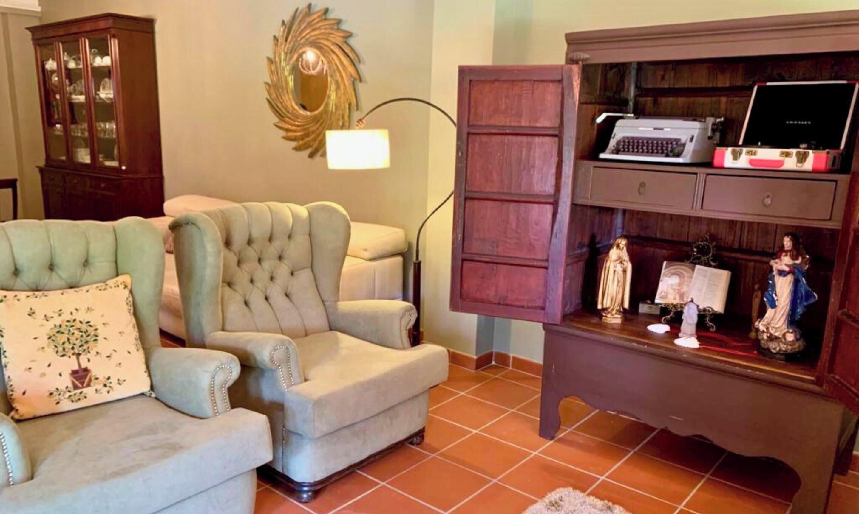 Un salon avec un fauteuil vert, un canapé beige, un meuble en bois sur lequel se trouve une machine à écrire, et un miroir décoratif.