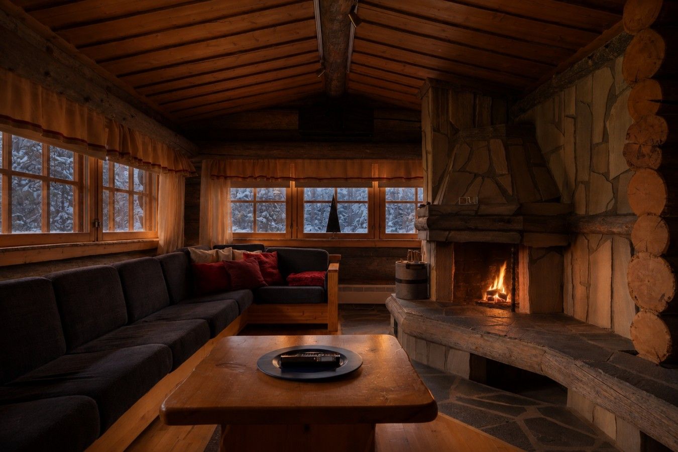 Interior acogedor de una cabaña con chimenea de piedra, mobiliario de madera y ventanas amplias.