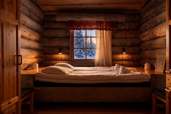 A log cabin room with a bed, two bedside lamps, a window with a view of snow-covered trees, and a red valance.