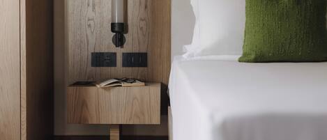 A hotel room with a wooden nightstand, a book, glasses, a lamp, and a bed with white bedding and a green pillow.