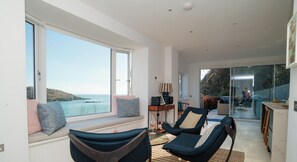A modern interior with a seating area by a large window overlooking the sea, a dining area, and a kitchen. - Luxury Estuary Views & Parking | Woodcot Cottage (Salcombe)