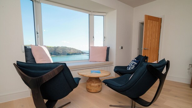 Ein Zimmer mit zwei Stühlen, die auf ein Fenster mit Blick auf ein Gewässer zeigen, einem Holztisch und einer Holztür.