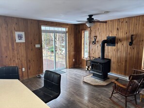 A room with wooden walls, a black upholstered chair, a wooden rocking chair, a black wood stove, and a ceiling fan.