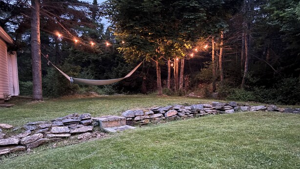 A grassy area with a stone wall, a hammock, and string lights in a wooded setting. - The Cottage at New Chelsea (New Chelsea)