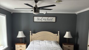A bedroom with a bed, two bedside tables with lamps, a ceiling fan, a window, and a wall sign. - The Cottage at New Chelsea (New Chelsea)