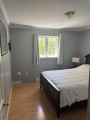 A bedroom with a wooden floor, a bed with white bedding, a window with curtains, and a ceiling light fixture.