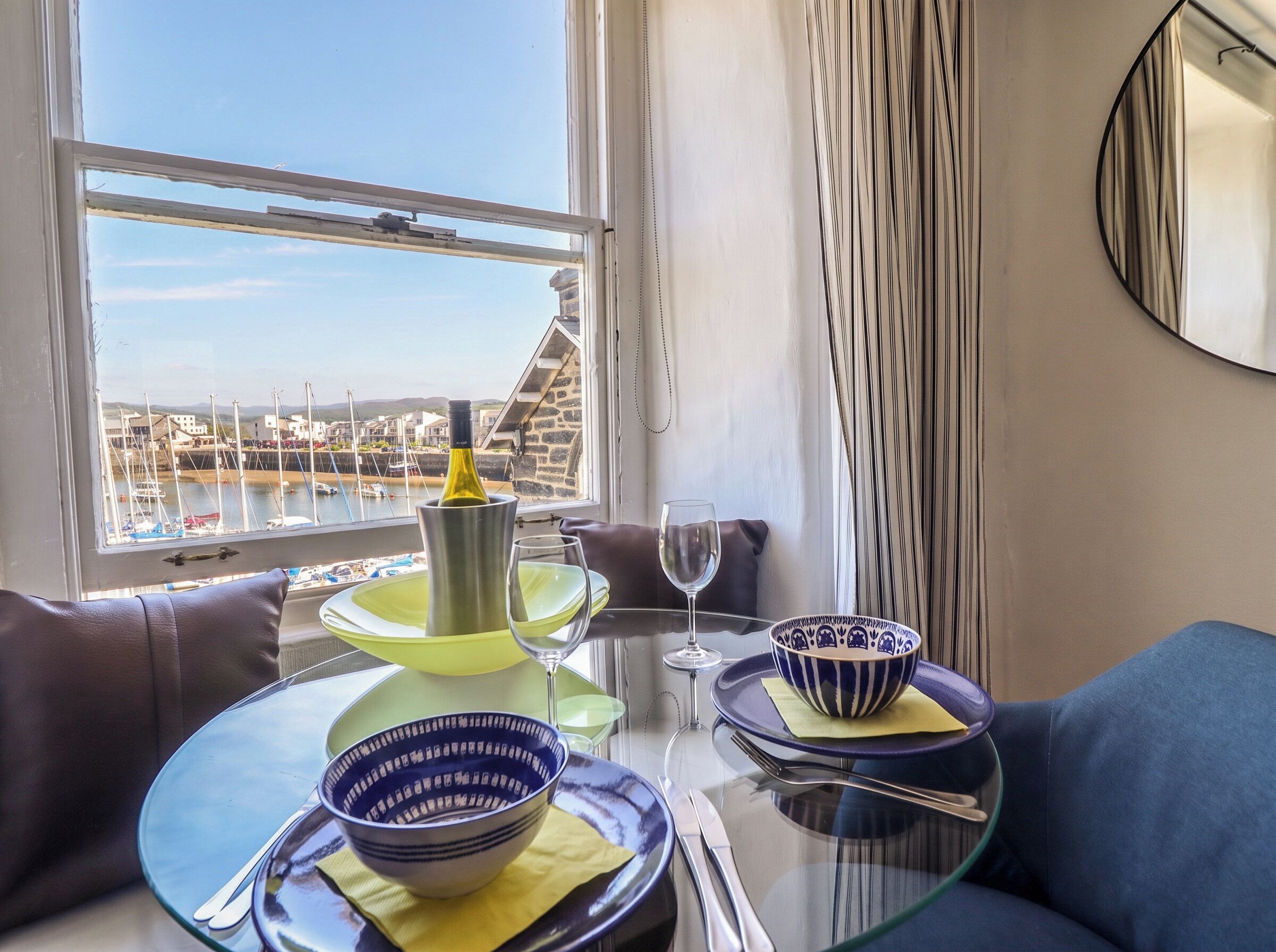 Een eethoek met een tafel gedekt voor twee personen, een wijnfles en uitzicht op een jachthaven.