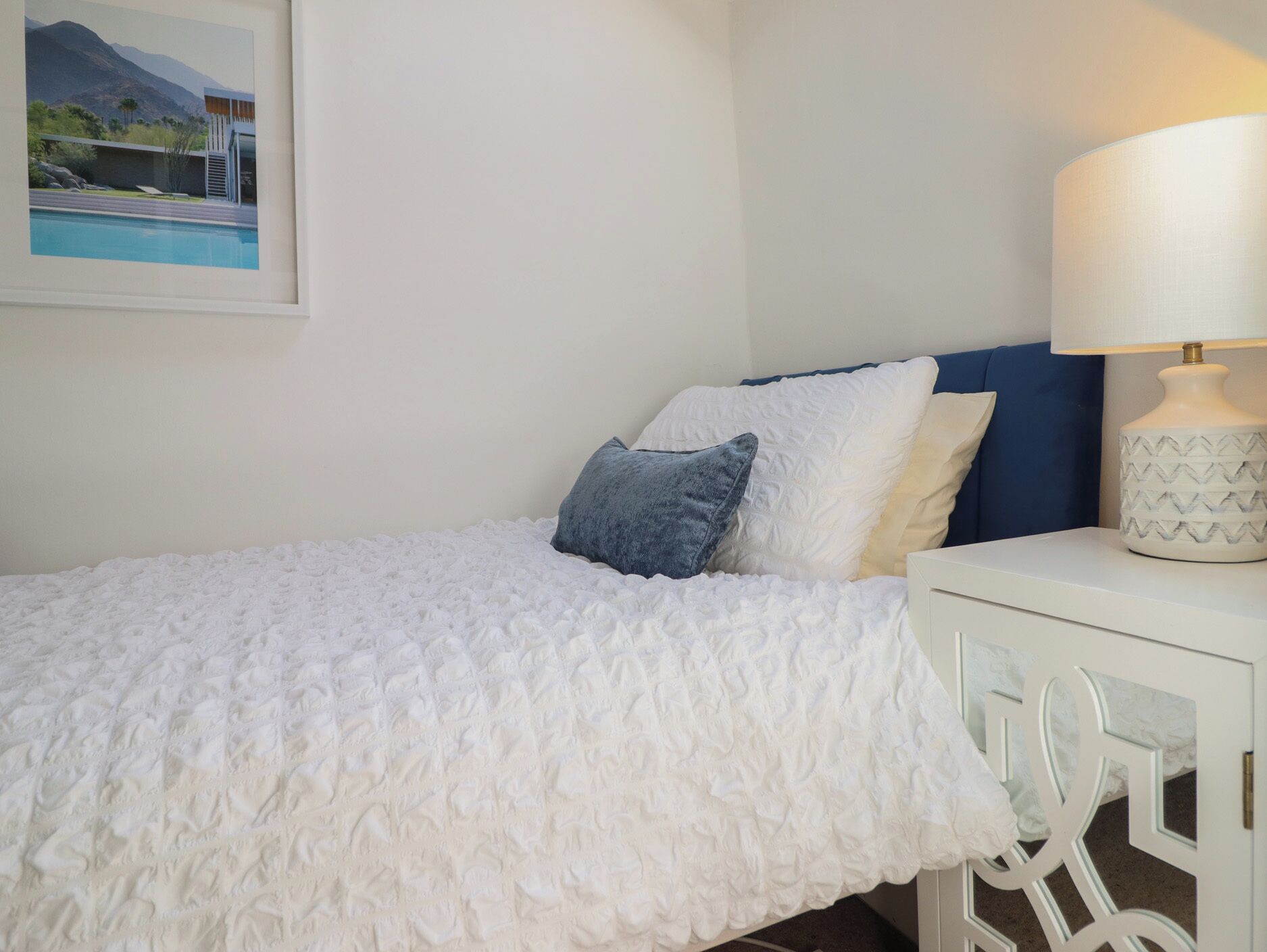 A neatly made bed with a textured white cover, a bedside table with a lamp, and a framed picture on the wall.