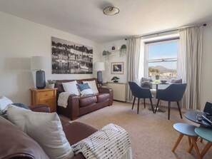 Ein Wohnzimmer mit braunem Ledersofa, einem Esstisch mit Stühlen, einem großen Fenster mit Vorhängen, einer Lampe und einem gerahmten Bild an der Wand.