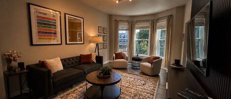A modern living room with a sofa, armchairs, a coffee table, and a large rug. There are framed pictures on the wall and a television on the right.