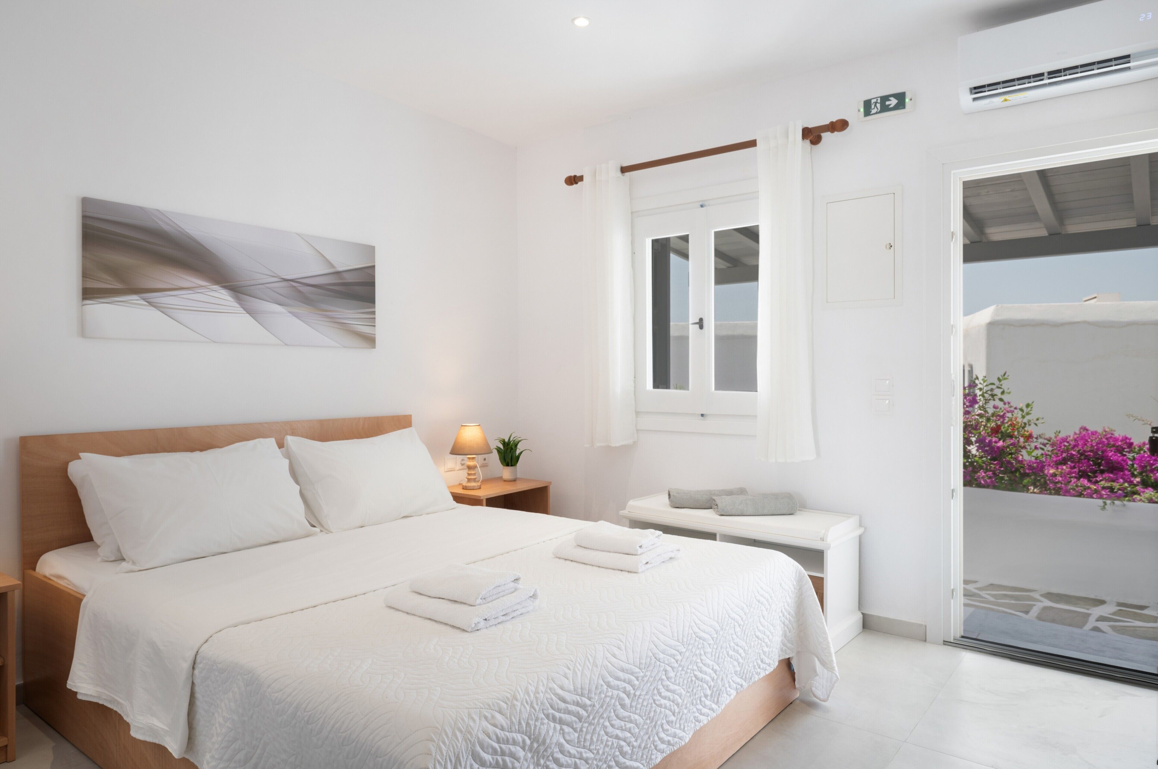 Ein Hotelzimmer mit einem Bett, einem Nachttisch mit Lampe, einem Fenster und einem Balkon mit Blumen.
