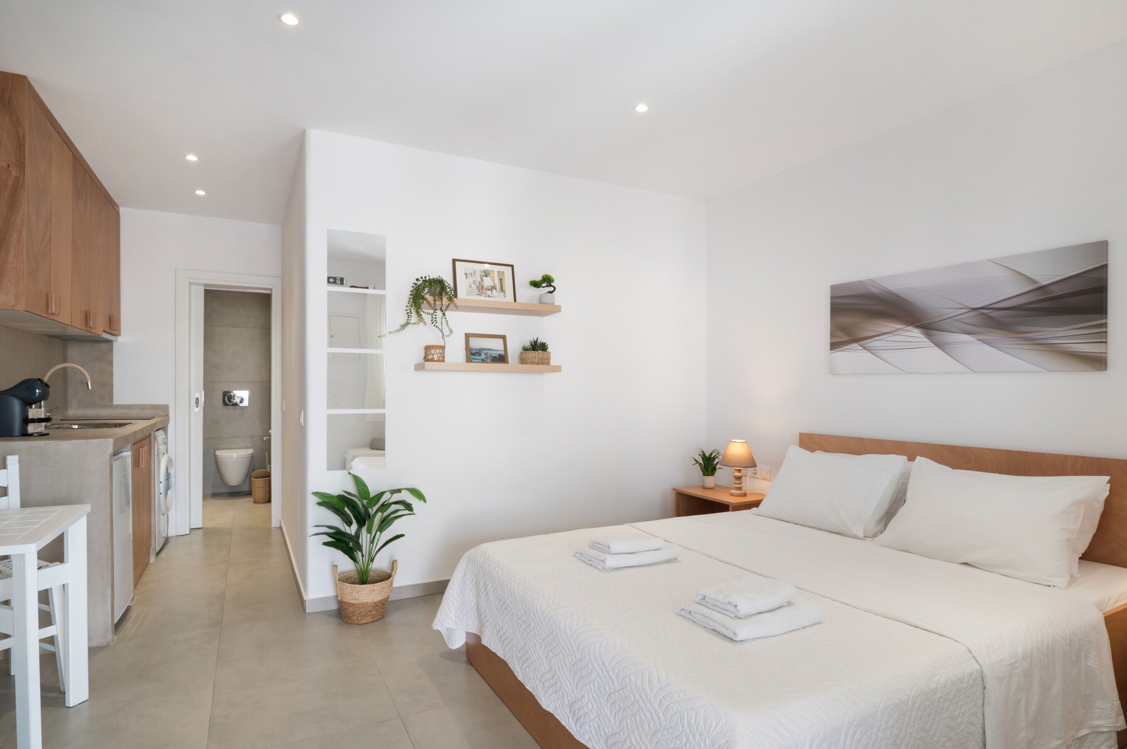 A modern bedroom with a large bed, a bedside table with a lamp, a wall-mounted shelf with decorative items, and a view of the bathroom.