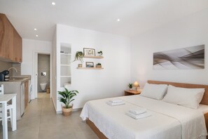 A modern bedroom with a large bed, a bedside table with a lamp, a wall-mounted shelf with decorative items, and a view of the bathroom. - Holiday Home 'Mykonos Serenity 1' with Shared Pool, Wi-Fi and Air Conditioning (Mykonos)