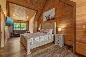 A bedroom with a wooden ceiling, a bed with patterned bedding, a nightstand with a lamp, and a large window with a view of trees. - Hilltop Dreamin': Life's Better Up Here (Sevierville)