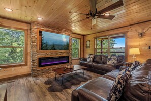 A cozy cabin interior with a stone fireplace, leather couches, and a large window offering a forest view. - Hilltop Dreamin': Life's Better Up Here (Sevierville)