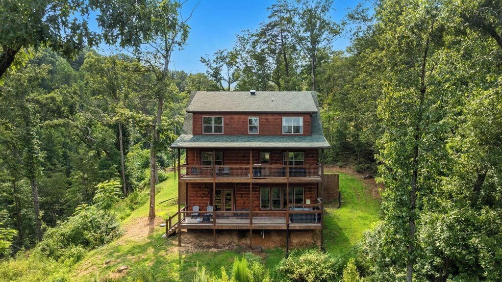 A two-story log cabin with a deck and multiple windows, surrounded by dense forest.