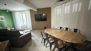 Zona pranzo con un tavolo rotondo in legno e sedie, una TV a schermo piatto montata a parete e una parete verde decorativa.