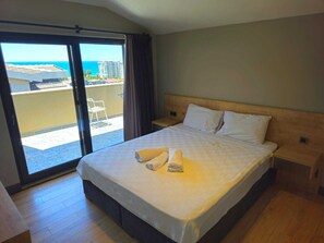 Une chambre d’hôtel avec un lit, une table de chevet, une fenêtre donnant sur des immeubles et la mer, et une porte coulissante donnant sur un balcon.