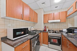 A compact kitchen with wooden cabinets, a microwave, a stove, and a dishwasher. - Bloomfields Studio in Manchester Tower (Dubai)