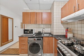 A compact kitchen with wooden cabinets, a washing machine, a microwave, a stove, and a sink. - Bloomfields Studio in Manchester Tower (Dubai)