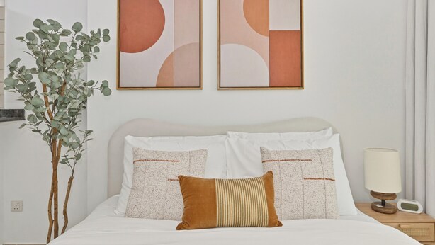 A hotel room with a bed, two wall-mounted abstract paintings, a potted plant, and a bedside table with a lamp and a radio. - Bloomfields Studio in Manchester Tower (Dubai)