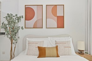 A hotel room with a bed, two wall-mounted abstract paintings, a potted plant, and a bedside table with a lamp and a radio. - Bloomfields Studio in Manchester Tower (Dubai)