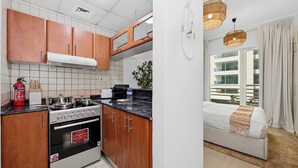 A kitchen with wooden cabinets, a stove, and a dining area with a sofa and curtains. - Bloomfields Studio in Manchester Tower (Dubai)