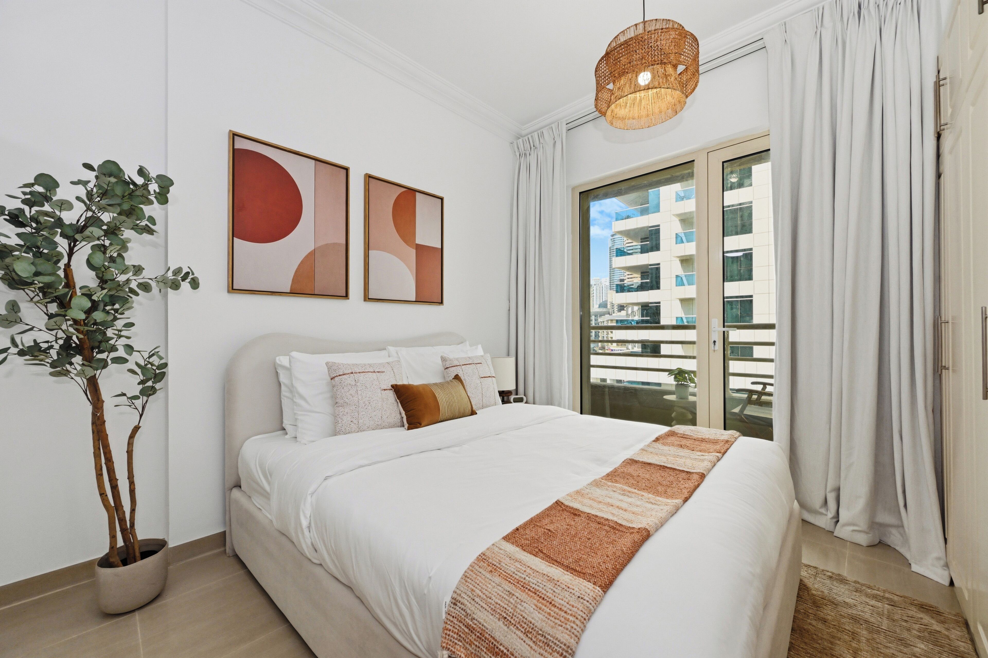 A modern bedroom with a large bed, a potted plant, abstract wall art, and a view of the city through a large window.