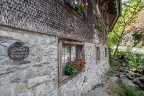 Un bâtiment en pierre doté d’un toit à pente traditionnelle, d’une fenêtre ornée de fleurs rouges et d’une plaque de pierre indiquant les coordonnées.