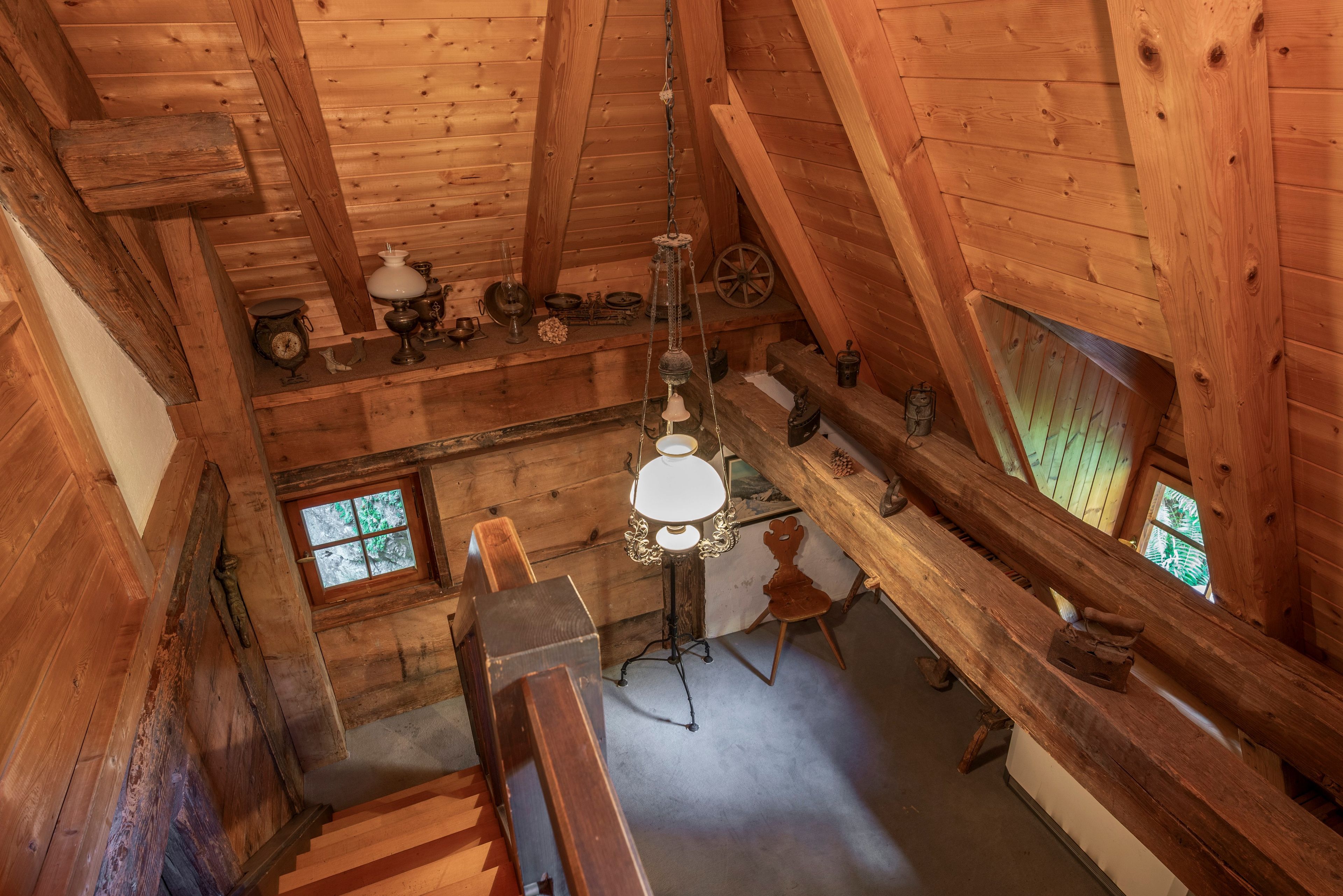 Ein Holzinnenraum mit schrägverdeckter Decke, einem Holzstuhl, einem Tisch mit einer Hängeleuchte und einem Fenster mit Blick auf Grünflächen.