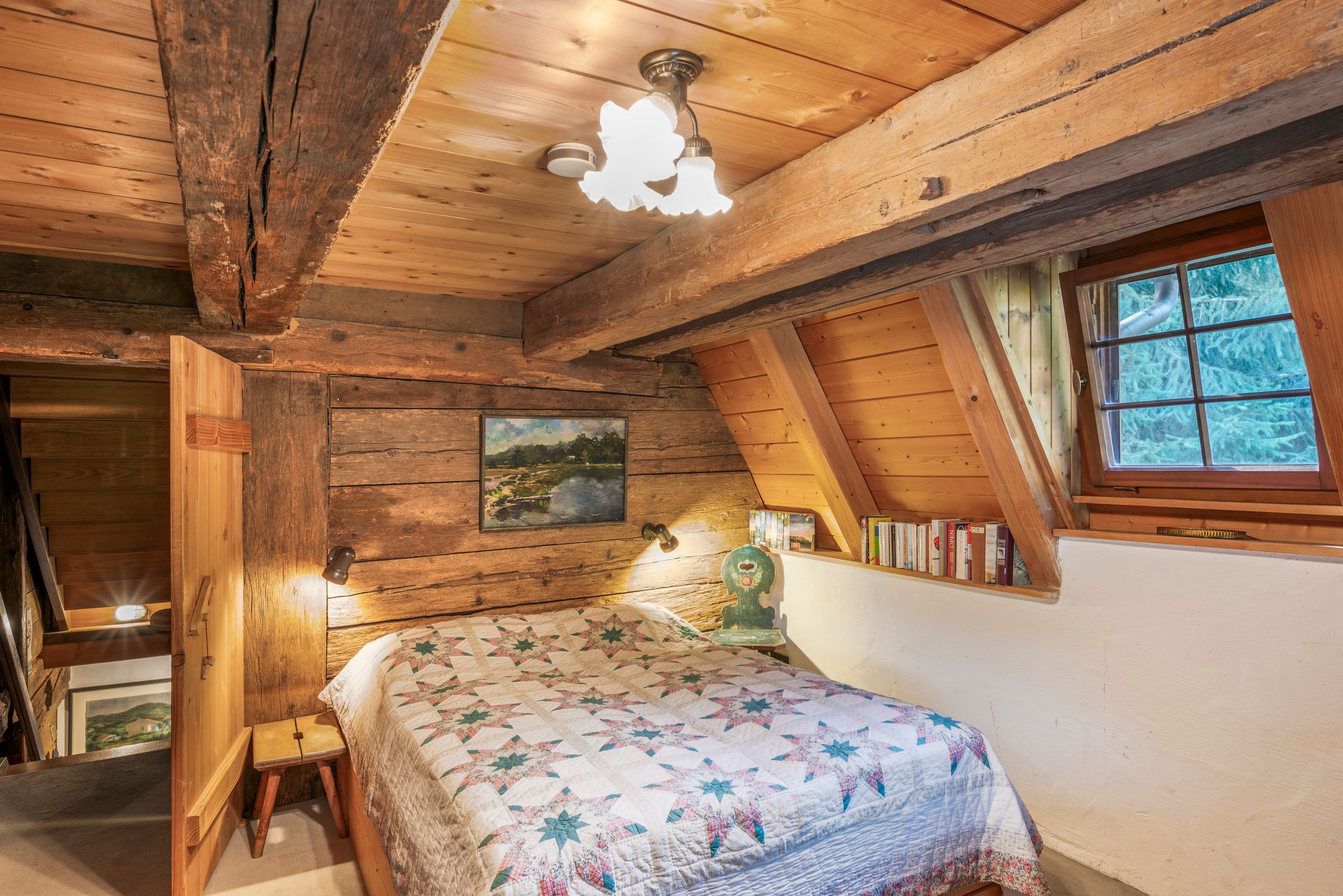 Une chambre mansardée chaleureuse avec un plafond en bois, un lit recouvert d’une courtepointe, une bibliothèque et une fenêtre donnant sur les arbres.