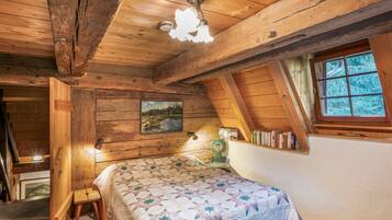 Quarto de sótão aconchegante com teto de madeira, cama com colcha, estante de livros e janela com vista para árvores.