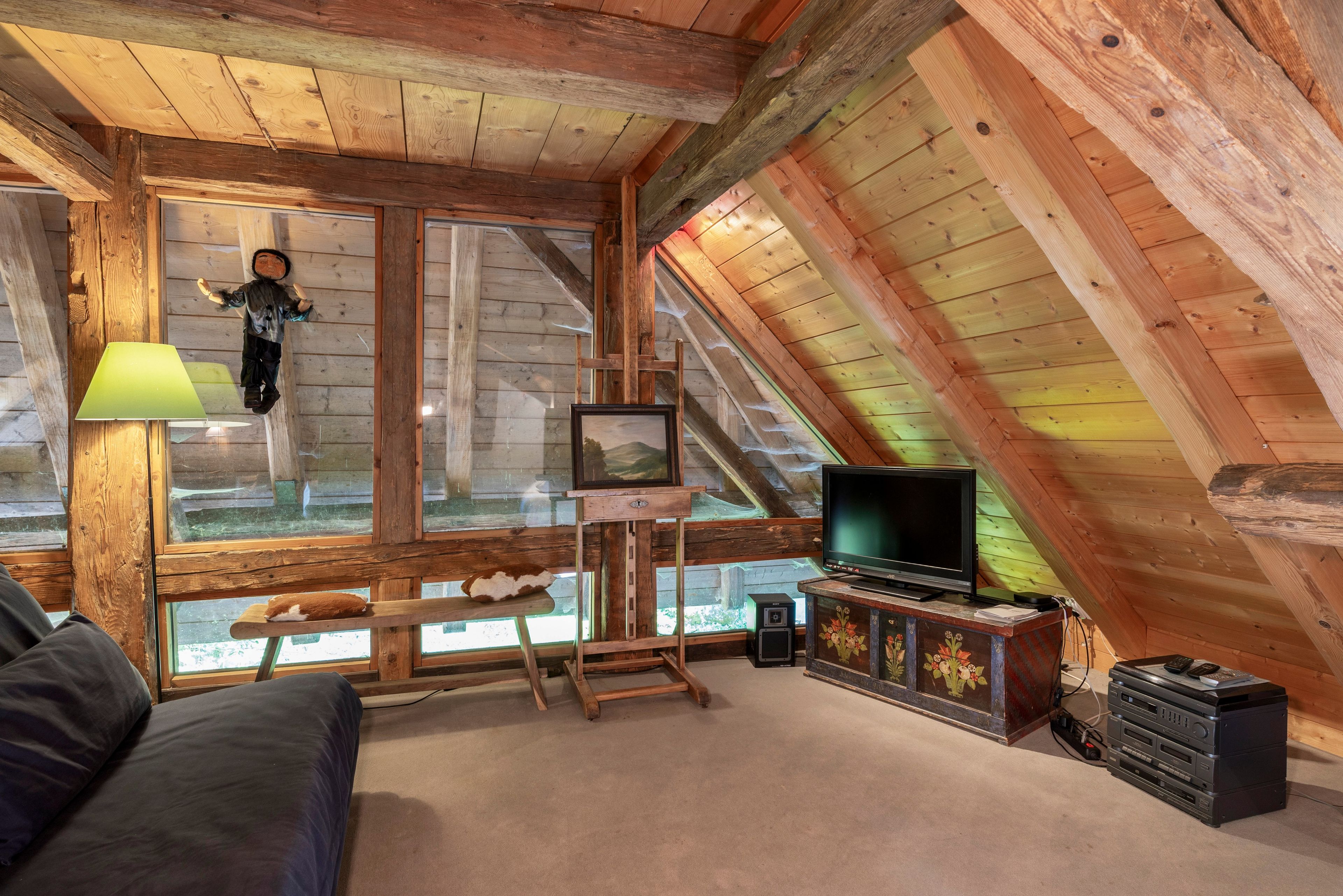 A cozy attic room with a wooden ceiling, a television, a painting on easel, and a plush sofa.
