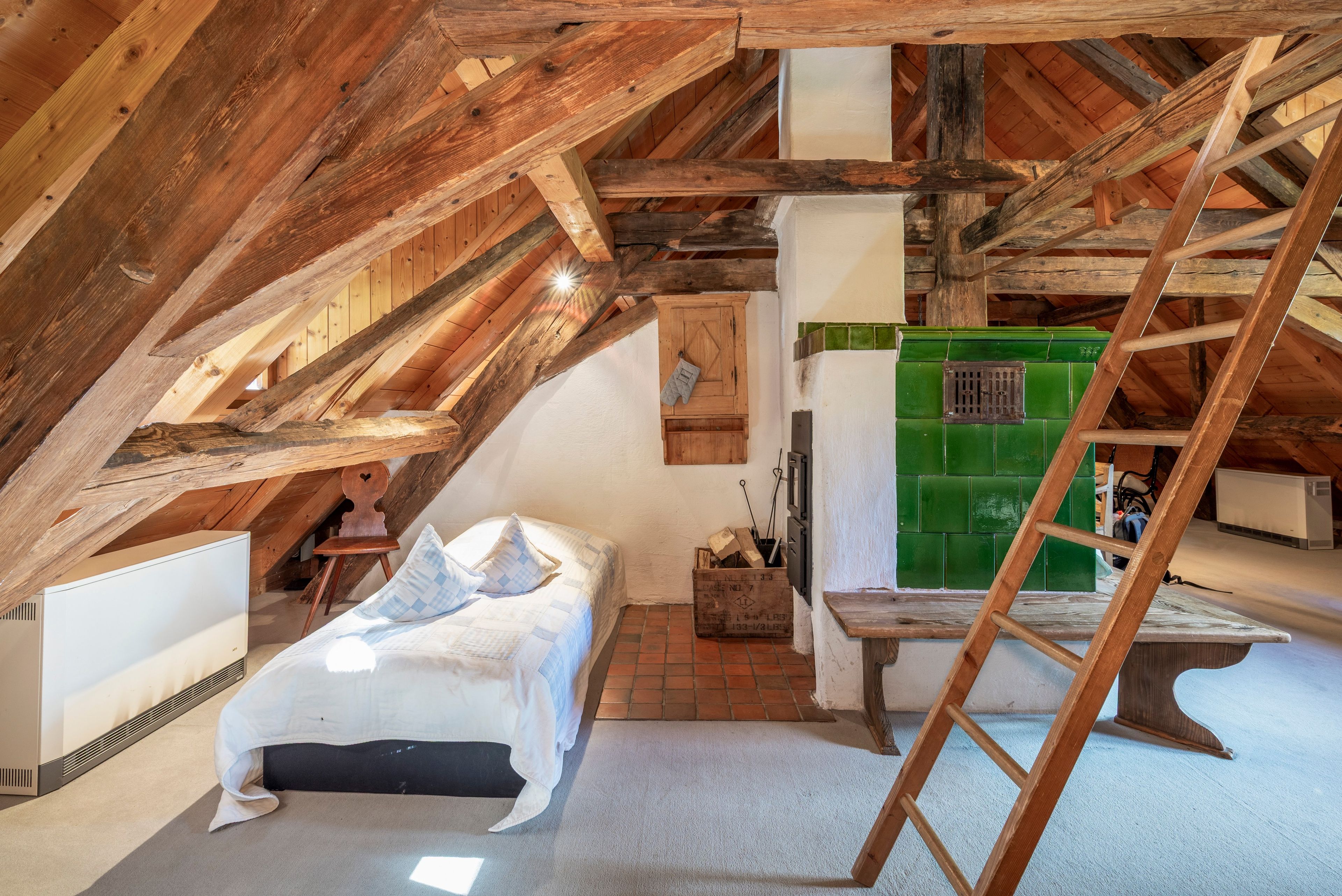 Ein Schlafzimmer im Spitzboden mit schrägem Dach, einer Holzleiter, einem Bett, einem kleinen Tisch und einem grünen Gerät.