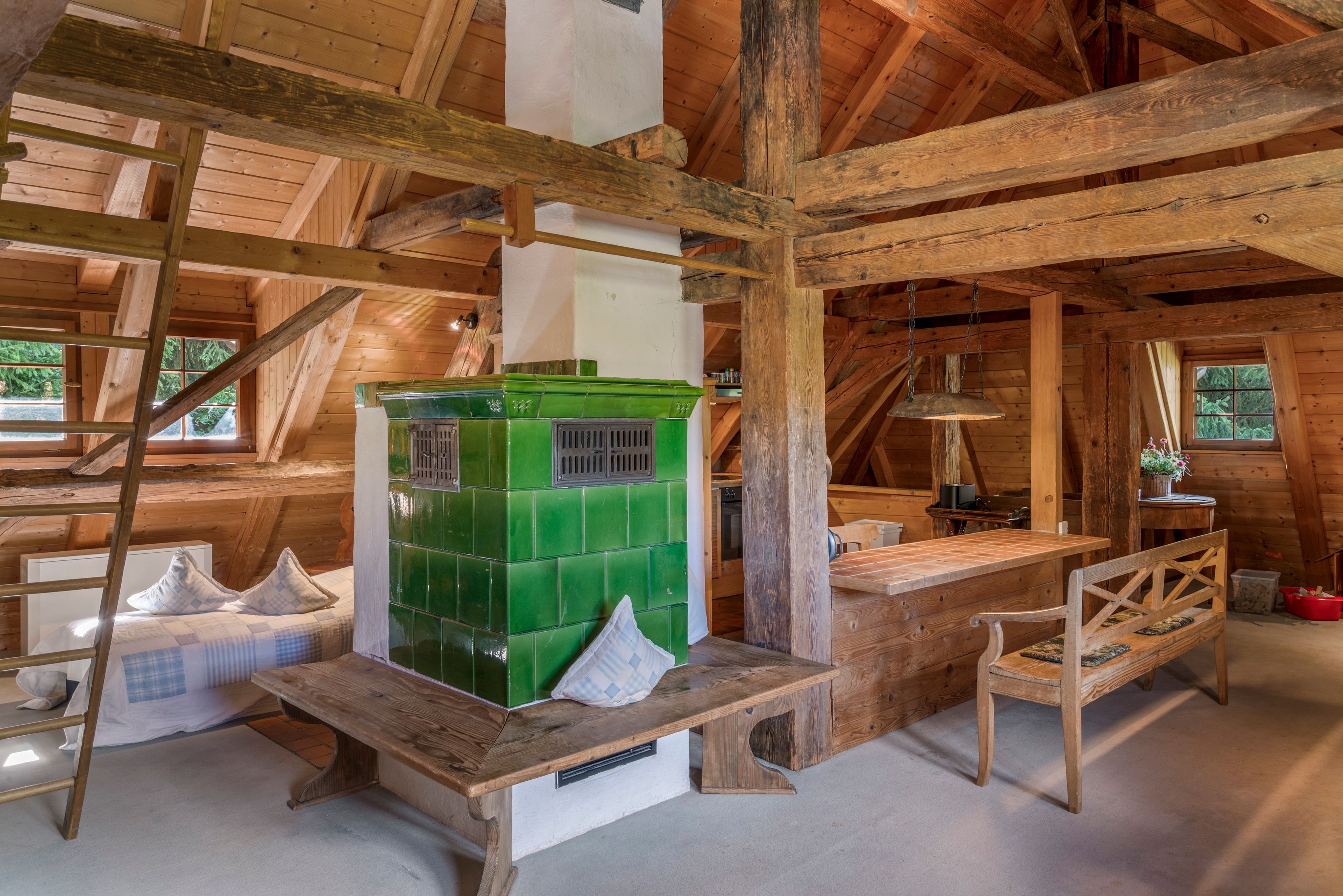 Un intérieur rustique en bois, avec un poêle en faïence verte, des bancs en bois et une échelle.