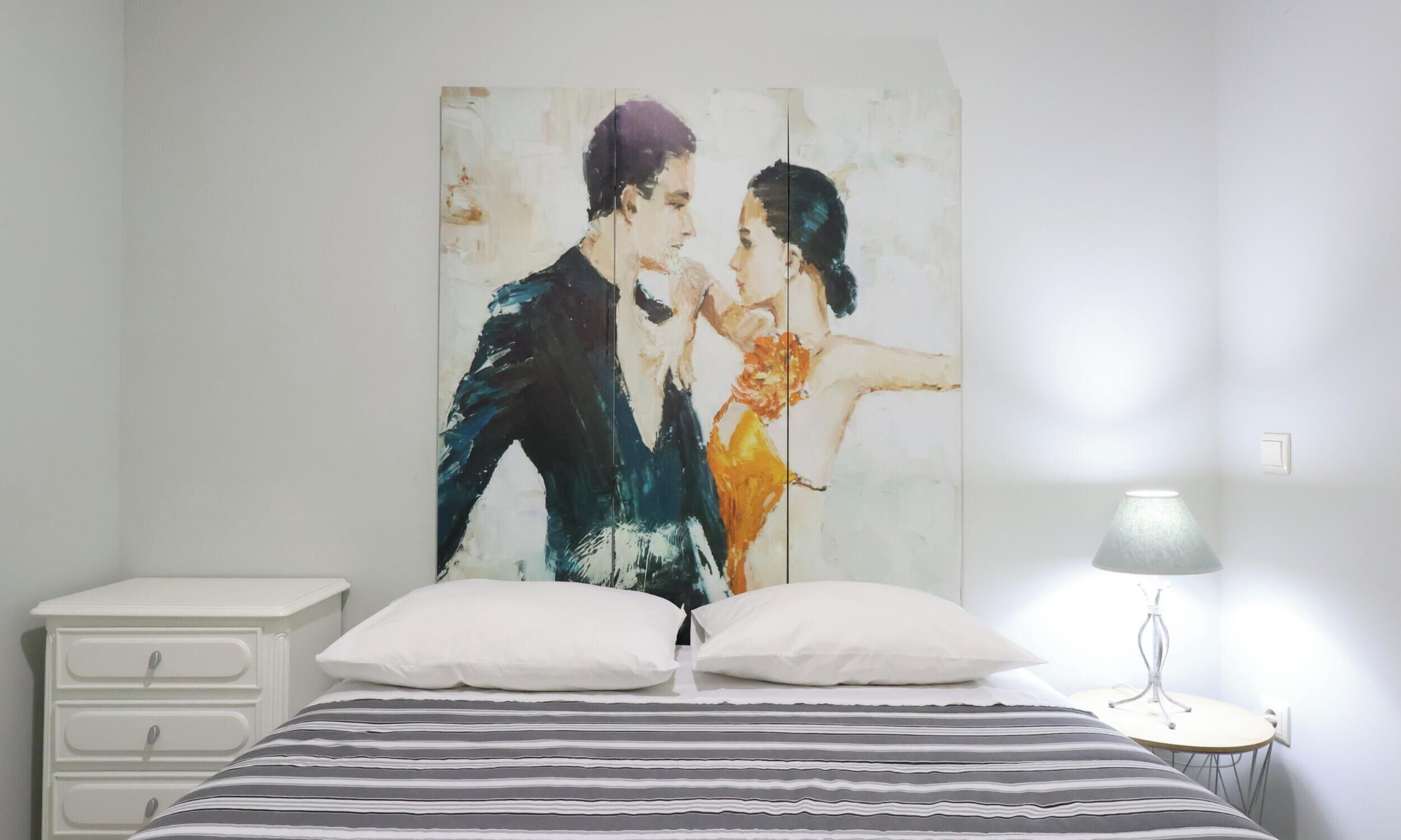 Ein Schlafzimmer mit einem Bett, zwei Kissen, einem Nachttisch, einer Lampe und einem Bild an der Wand.