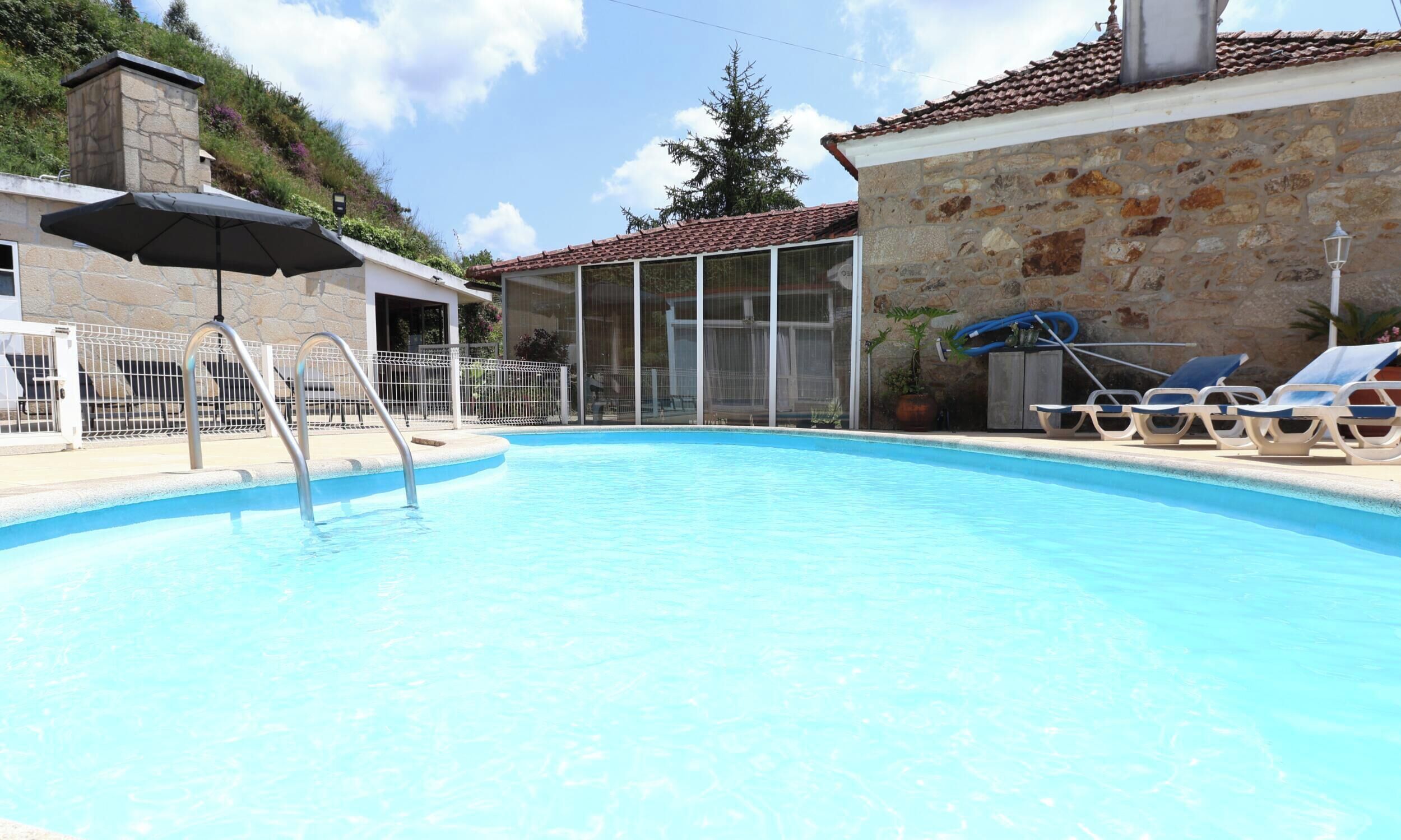 A swimming pool with a metal ladder, lounge chairs, and a covered area with an umbrella.