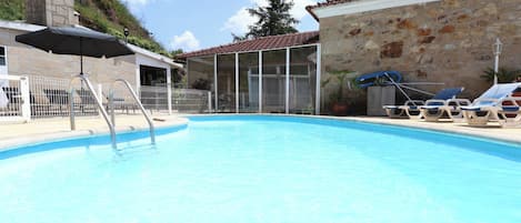 A swimming pool with a metal ladder, lounge chairs, and a covered area with an umbrella.