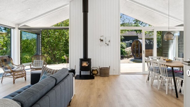 Un vaste espace de vie doté d’un haut plafond, d’un poêle à bois central, d’un canapé gris, de fauteuils en osier et d’un coin repas avec des chaises blanches et une table en bois.