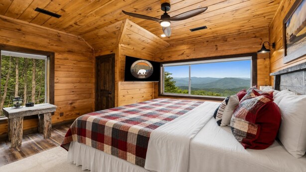 Ein Schlafzimmer in einer Holzhütte mit einem großen Bett, Blick auf die Berge und einem Deckenventilator.