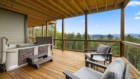Een overdekt terras met een stenen barbecue, houten meubilair en uitzicht op een bos en bergen.
