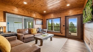 L’intérieur chaleureux d’un chalet avec un plafond en bois, un canapé en cuir, une table basse et une grande fenêtre donnant sur le paysage extérieur.