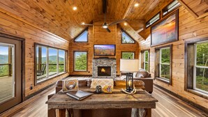 A cozy cabin interior with a fireplace, wooden walls, large windows, and a ceiling fan. - Thunderhead Mountain Lodge- Amazing Views, Amazing Private Pool & Game Room (Sevierville)