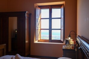 A bedroom with a large window, a bedside table with a lamp, and a wooden wardrobe.